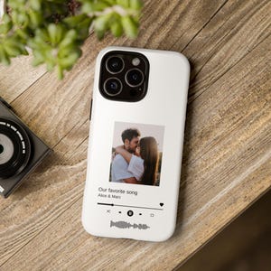 Könnte beinhalten: Weißes Handycase mit einem Foto eines sich umarmenden Paares. Das Case zeigt den Text "Our favorite song" und eine Musikplayer-Oberfläche. Das Handy liegt auf einer Holzoberfläche.