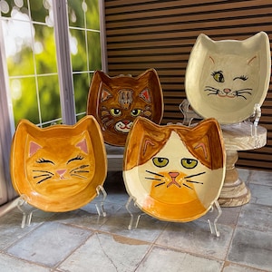 May include: Four ceramic cat-shaped dishes with painted faces. The dishes are in shades of orange, brown, and cream, each with a unique cat expression. They are displayed on clear stands, showcasing their whimsical design.