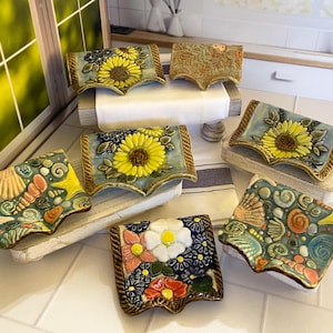 May include: A collection of six rectangular ceramic spoon rests with curved edges. Three feature yellow sunflowers on blue backgrounds, two display colorful seashells and starfish on teal, and one shows white and red flowers on a dark blue base.