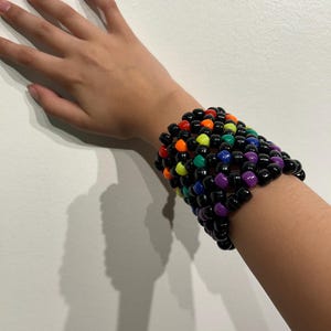 May include: A rainbow kandi bracelet made with black and colorful beads. The bracelet is worn on a person's wrist.