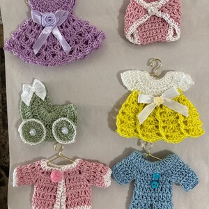 May include: Six crocheted baby outfits, including a purple dress, a pink nappy, a yellow dress, a green pram, a pink cardigan, and a blue romper.