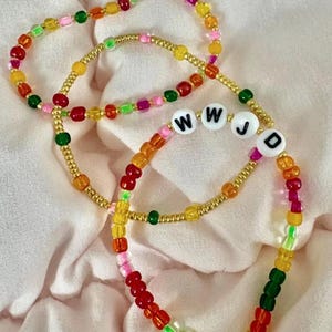 Könnte beinhalten: Zwei elastische Armbänder aus kleinen, bunten, durchscheinenden Perlen in Rot, Orange, Gelb, Grün und Pink, durchsetzt mit metallisch goldenen Perlen. Ein Armband enthält weiße, runde Perlen mit den schwarzen Buchstaben W, W, J und D.