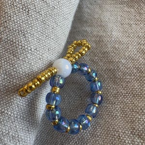 May include: A beaded ring with a blue iridescent band and gold accents. A white bead and a gold beaded extension are attached to the ring. The ring is set against a neutral gray background.