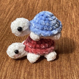 May include: Two crocheted plush turtle toys stacked on top of each other. The bottom turtle is white with a pink shell and the top turtle is white with a blue shell.