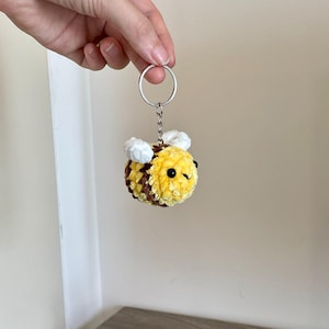 May include: A crocheted bee keyring made with yellow, brown, and white yarn. The bee has black eyes and is attached to a silver keyring. The keyring is held up by a hand.