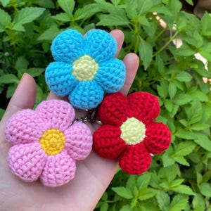 May include: Three handmade crochet flower keyrings in blue, pink, and red, each with a yellow centre. The flowers are held against green foliage, showcasing the vibrant colours and textured details of the yarn.