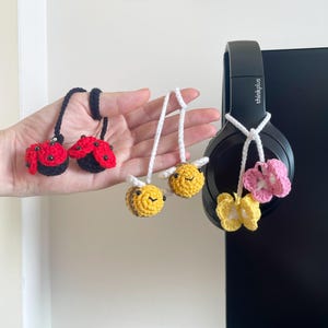 May include: Crocheted ladybug, bee, and flower charms. The ladybugs are red and black, the bees are yellow and brown, and the flowers are pink and yellow. The charms are attached to white cords.