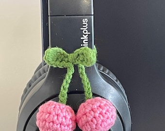 Pode incluir: Fones de ouvido pretos adornados com um laço verde de crochê e duas cerejas rosa de crochê. Os fones de ouvido exibem a palavra "inkplus".