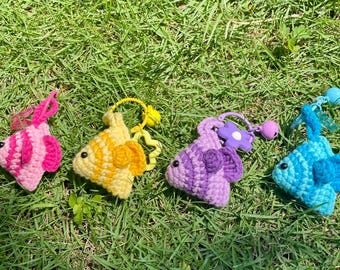 May include: Four colorful crochet fish keychains in pink, yellow, purple, and blue, each with a different charm. The fish are lying on green grass, with the sun shining on them.