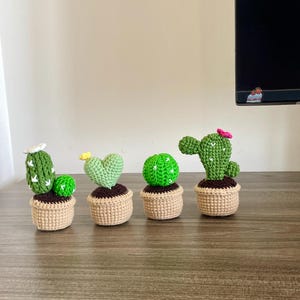 Op de afbeelding: Vier handgemaakte gehaakte cactusplanten in kleine beige potten. De cactussen zijn in verschillende groentinten, met kleine witte, gele en roze bloemaccenten. De potten zijn gemaakt van beige garen en staan op een houten oppervlak.