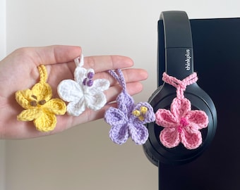 May include: Four crochet flower charms in yellow, white, purple, and pink. The charms are held in a hand and one is attached to a black headset. The headset has the text "thinkplus" on it.