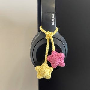 Peut inclure: Casque audio noir avec un cordon crocheté jaune et deux breloques en forme d'étoile, une rose et une jaune. Le casque est suspendu à une surface noire. La marque "thinkplus" est visible.