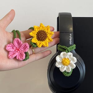 Op de afbeelding: Gehaakte bloemenaccessoires in roze, geel en wit met groene accenten. Een zwarte headset heeft een witte en gele madelief bevestigd. De andere bloemen zijn een roze bloem en een gele zonnebloem.