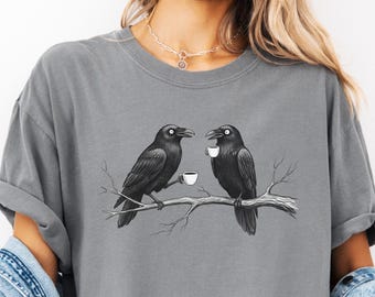 May include: Grey t-shirt featuring a black and white illustration of two crows perched on a branch, each holding a small teacup. The shirt is a comfortable, casual style.