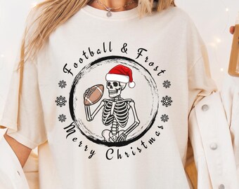 May include: Ivory-colored t-shirt featuring a circular graphic with a skeleton wearing a Santa hat and holding a football. The text reads "Football & Frost" and "Merry Christmas" around the skeleton. The shirt has short sleeves.
