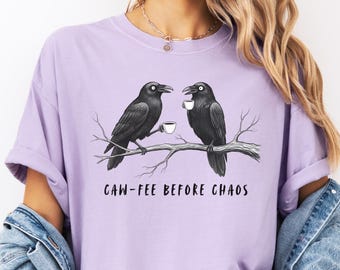 May include: A light purple t-shirt featuring a graphic of two black crows perched on a branch, each holding a coffee cup. The text "CAW-FEE BEFORE CHAOS" is printed below the illustration. The shirt is part of a clothing collection.
