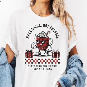 May include: White t-shirt with a graphic of a cocoa mug wearing sunglasses and giving a thumbs up. The text reads "MAKE COCOA, NOT EXCUSES" and "SLEIGHING GOALS ONE SIP AT A TIME". Two gift boxes are on either side of the mug.