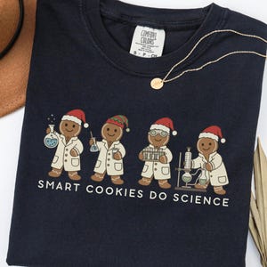 May include: Black crew neck t-shirt featuring a graphic of four gingerbread cookies in lab coats and Santa hats, conducting science experiments. The text "SMART COOKIES DO SCIENCE" is printed below.