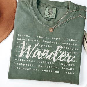 May include: A moss green t-shirt with the word "Wander" in white script. The shirt features a list of travel-related words. A gold necklace with a pendant rests on the shirt. The shirt is labeled "Comfort Colors".