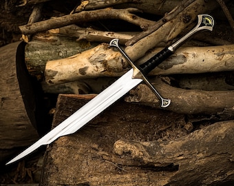May include: A silver sword with a double-edged blade, a black leather-wrapped handle, and a decorative hilt. The sword rests on a pile of wooden logs, showcasing its craftsmanship and design. The hilt features a gold and silver design.