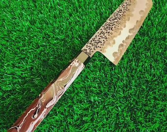 May include: A kitchen knife featuring a hammered metal blade with a visible wavy temper line and a polished edge. The handle is constructed from a brown, white, and gold marbled resin material, connected to the blade by a dark metal bolster.