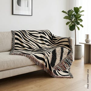 May include: A black and beige zebra-striped throw blanket with fringe detail is draped over a beige sofa. The blanket's pattern is a bold, repeating design. The close-up shows the woven texture and fringe.