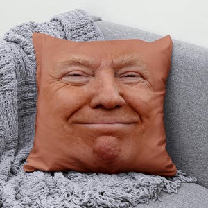 May include: A square pillow featuring a close-up of a smiling face with fair skin and reddish-blonde hair. The pillow is a light peach color and rests on a gray couch with a gray knitted blanket.