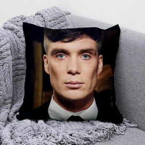 May include: Square throw pillow featuring a portrait of a man with blue eyes, dark hair, and a white collared shirt. The pillow is black and rests on a gray sofa with a gray knitted throw blanket.