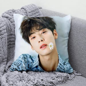 May include: A cushion with a portrait of a person with dark hair and a daisy in their mouth. The person is wearing a blue shirt with white text. The cushion is on a grey sofa with a grey knitted blanket.