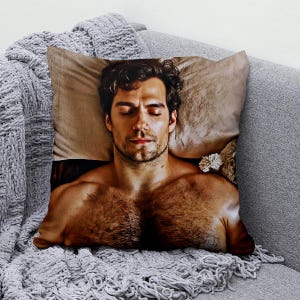 May include: A cushion with a printed image of a man with closed eyes, lying on a pillow. The pillow has a beige background and is placed on a grey sofa with a grey blanket.
