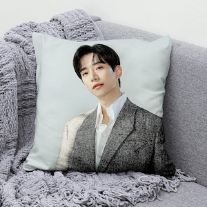 May include: Square decorative pillow featuring a portrait of a person wearing a white shirt and a gray tweed blazer. The pillow is on a gray sofa with a gray knitted throw blanket.