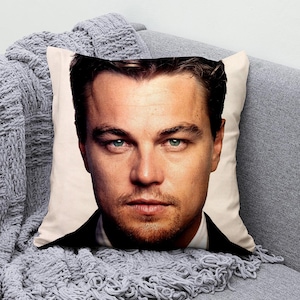 May include: Square decorative pillow featuring a close-up portrait of a man with light skin, blue-green eyes, and a short beard. The pillow is on a gray couch with a gray knitted throw blanket.