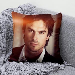 May include: Square decorative pillow featuring a portrait of a man with dark hair and light eyes. The pillow has a black suit and white shirt. The pillow is on a gray couch with a gray knitted throw blanket.