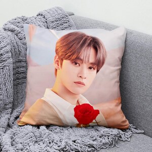 May include: Square decorative pillow featuring a portrait of a person holding a red rose. The person has brown hair and is wearing a white shirt. The pillow is on a gray couch with a gray knitted blanket.