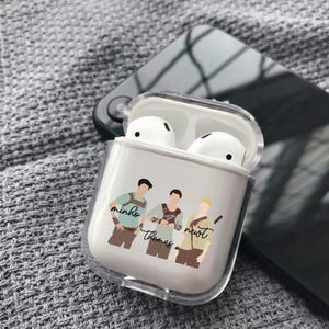 May include: A clear protective case for wireless earbuds containing a pair of white earbuds. The front of the case features a graphic illustration of three figures with the names Minho, Thomas, and Newt printed in black cursive script below them.