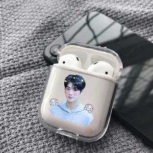 Puede incluir: Una funda protectora de plástico transparente para auriculares inalámbricos con un gráfico impreso de una persona de pelo oscuro con una sudadera azul claro, decorada con pequeños personajes de dibujos animados y un efecto de corona brillante.