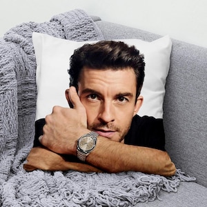May include: White square pillow featuring a portrait of a man with dark hair and a silver wristwatch. The man is resting his arm on a gray knitted blanket. The pillow is on a gray sofa.
