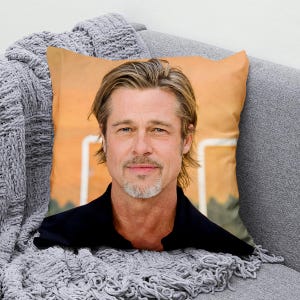 Könnte beinhalten: Quadratisches Kissen mit einem Porträt eines Mannes mit blonden Haaren und einem Spitzbart. Das Kissen hat einen pfirsichfarbenen Hintergrund und liegt auf einem grauen Sofa mit einer grauen Strickdecke.