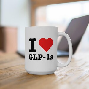 Puede incluir: Taza de café de cerámica blanca con un diseño en negro y rojo. La taza tiene el texto "I love GLP-1s" con un corazón rojo.