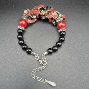 May include: A beaded bracelet featuring black and red beads, and small red and green flower accents. The bracelet has a silver-toned clasp and extender chain. The bracelet is designed with a mix of textures and colors.