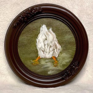 Op de afbeelding: Een kleine, ronde, ingelijste schildering van een witte eend met oranje voeten. De eend is geschilderd in een textuurstijl, met de achterkant en voeten zichtbaar. De lijst is donkerbruin met bloemmotieven.