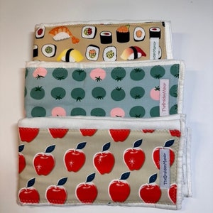 May include: Three rectangular cloths with different patterns. The top cloth has a sushi design on a tan background. The middle cloth has a green and pink tomato pattern on a blue background. The bottom cloth has a red apple pattern.