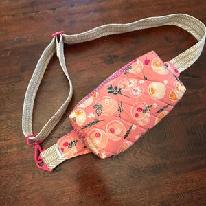 May include: A pink quilted fanny pack with a fruit pattern, featuring peaches and oranges. The bag has a beige strap with pink hardware. The fanny pack is lying on a dark wooden surface.