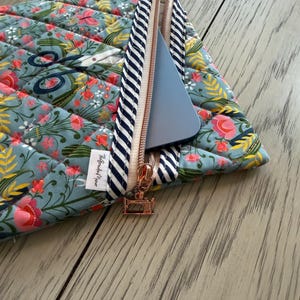 May include: Quilted floral laptop sleeve in teal, pink, and yellow. The sleeve has a diagonal quilted pattern and a navy and white striped zipper. A rose gold zipper pull and a small white tag with black text are visible.