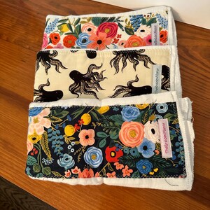 May include: Three folded fabric cloths with different patterns. One has a floral design with pink, orange, and blue flowers. Another features black octopus illustrations on a cream background. The third cloth has a dark blue background with colorful flowers.