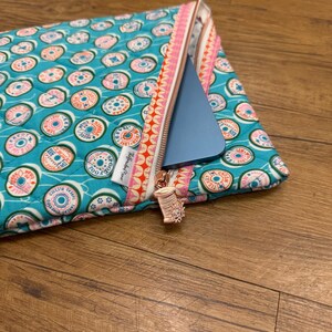 May include: A quilted fabric pouch in teal featuring a repeating pattern of colorful thread spools. The pouch has a zipper closure with a metallic spool-shaped charm and a patterned trim. A blue electronic tablet is partially inside the pouch.