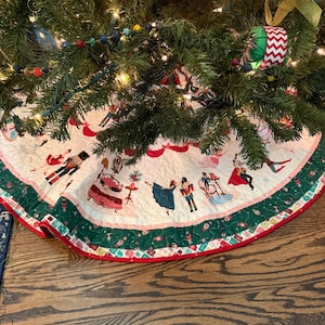 May include: A circular Christmas tree skirt featuring a white quilted fabric base decorated with colorful illustrations of nutcrackers, ballerinas, and holiday scenes. The border is a wide band of dark green fabric with a red and white geometric trim.