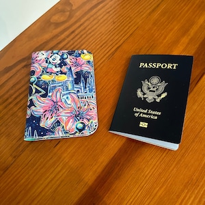 May include: A passport and a passport holder are displayed on a wooden surface. The passport is black with gold lettering and the words "PASSPORT" and "United States of America." The passport holder has a colorful floral and cartoon design.