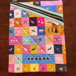 May include: A quilted fabric pouch with a colorful patchwork design. The squares feature various whimsical illustrations, including cats, birds, and sunglasses. A black zipper with a mint green trim runs diagonally across the top. The pouch is likely used for carrying small items.