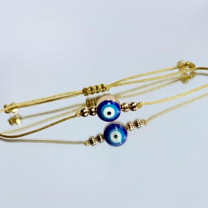 May include: Two gold-colored string bracelets with adjustable knots. Each bracelet features a blue and white evil eye bead, along with smaller gold-colored beads. The bracelets are displayed on a reflective surface.